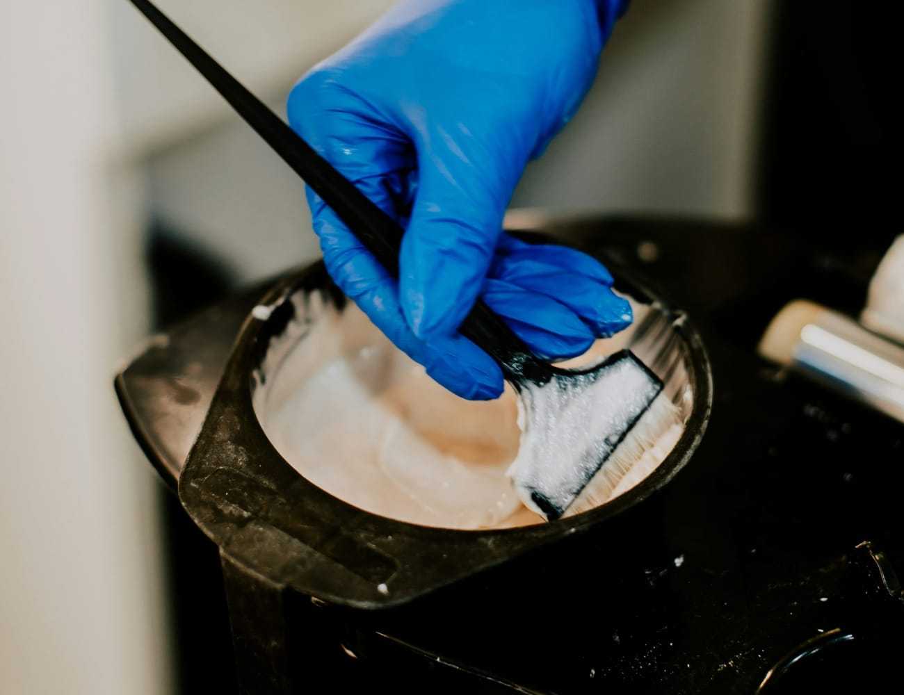 Gloved hand mixing hair dye in a bowl with a brush.